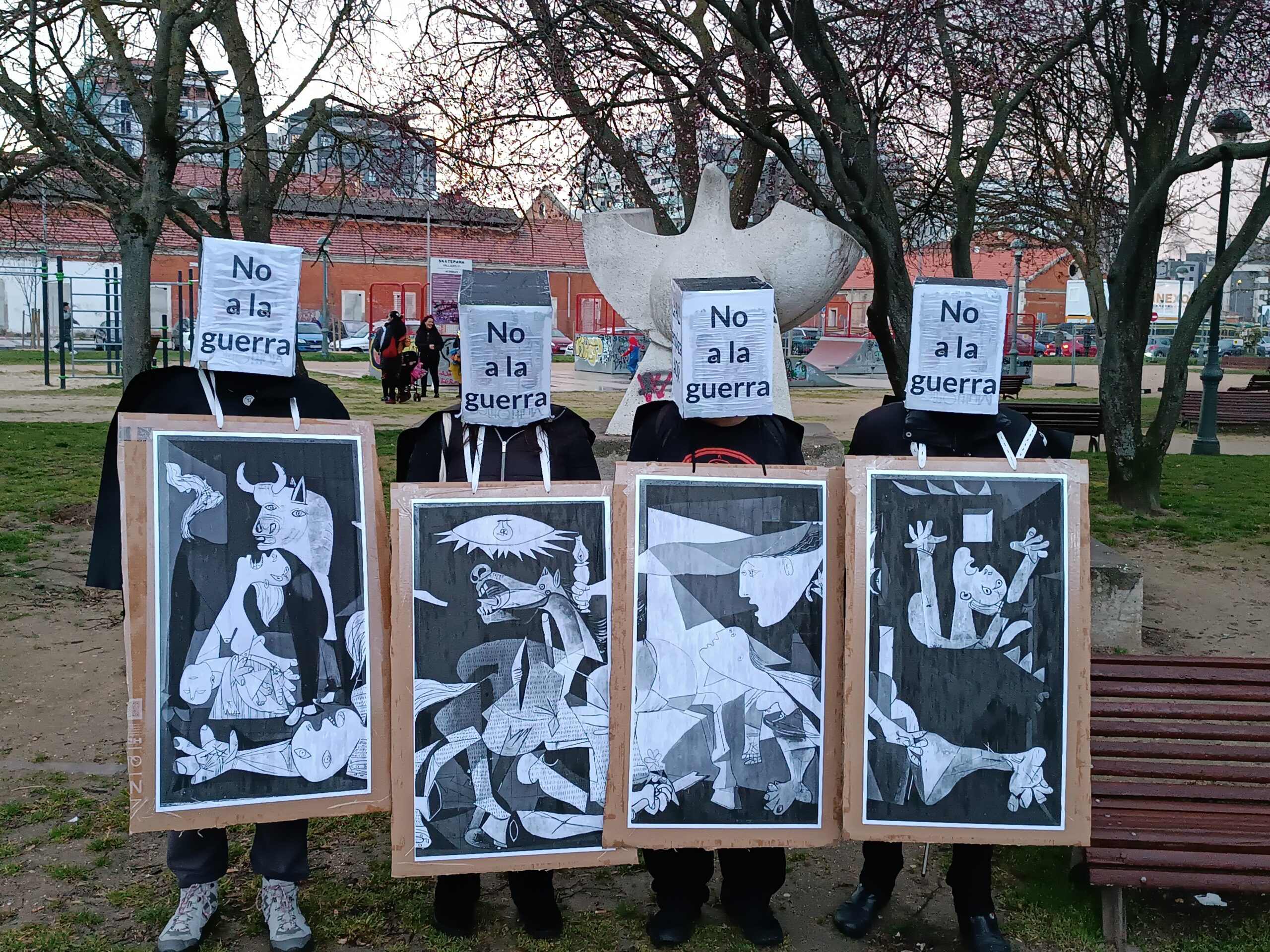 Cuatro personas, cada una portando un fragmento del Guernica en un gran cartón adosado a su cuerto. Al fondo una estatua de una paloma en el parque de la Paz.