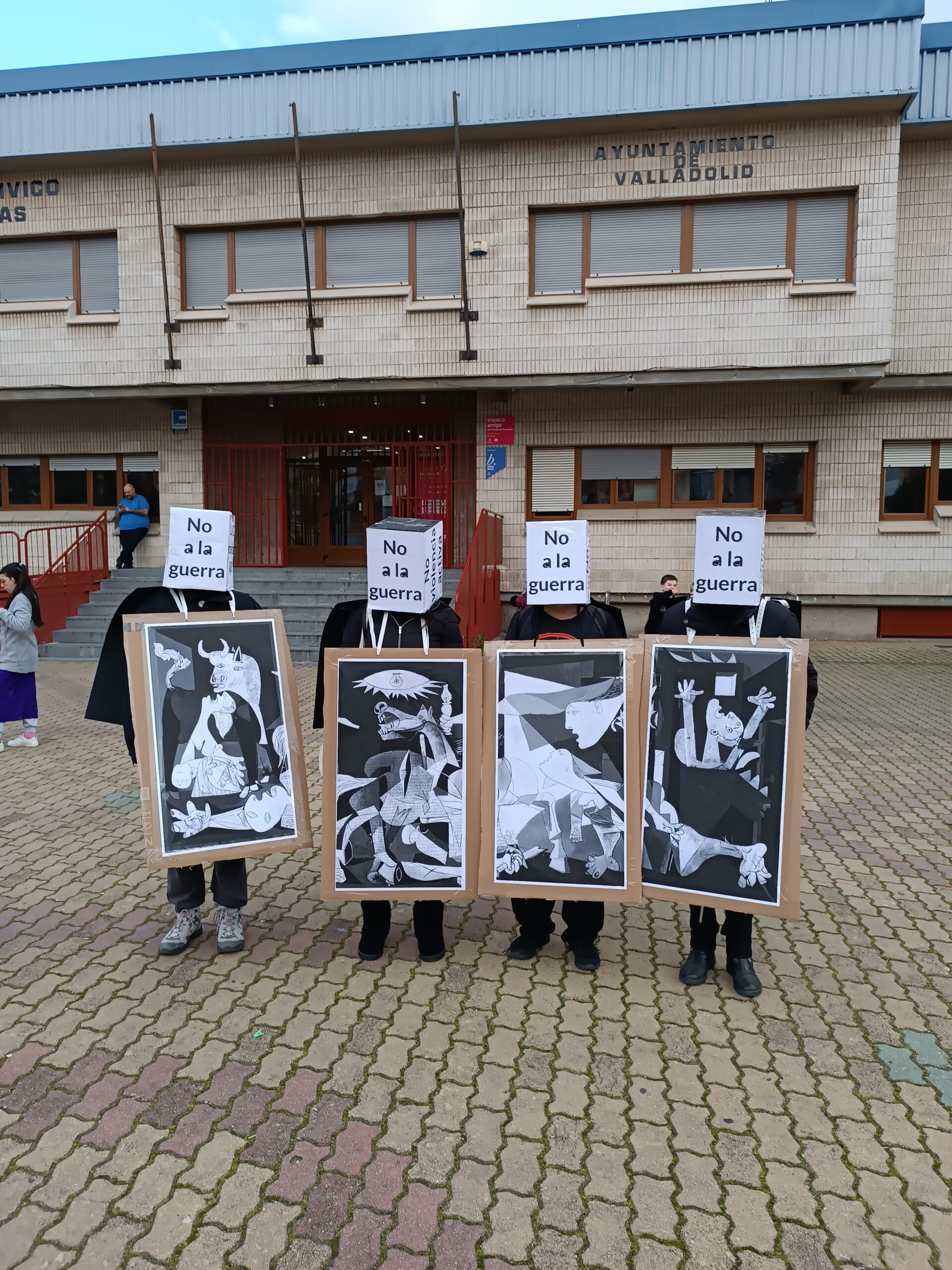 Cuatro personas, cada una portando un fragmento del Guernica en un gran cartón adosado a su cuerto. Al fondo la fachade del centro cívico Delicias.